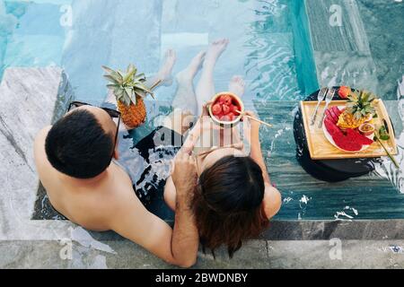 Junges Paar, das sich im Swimmingpool des Spa-Resorts entspannt und ein köstliches Frühstück genießt Stockfoto