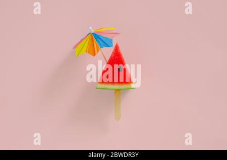 Rote Scheibe Wassermelone Design als Eis mit Stick, die Cocktail-Regenschirm auf rosa Hintergrund haben. Minimales Sommerobstkonzept. Stockfoto