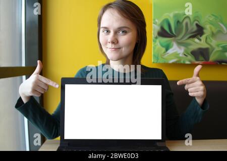 Ein schönes Mädchen sitzt in einem Café an einem Tisch vor dem Hintergrund einer gelben Wand und zeigt ihre Finger auf dem Bildschirm des Laptops. Der Computerbildschirm hat einen weißen rechteckigen Raum für Text. Design-Layout. Stockfoto