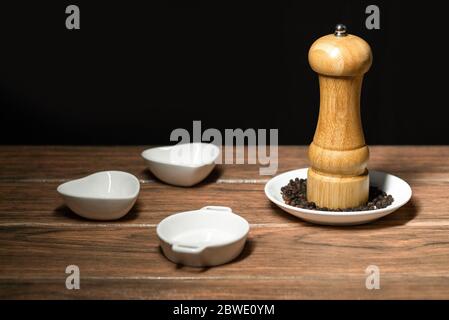 Schwarze Pfeffermühle mit ganzen Pfefferkörnern auf einer weißen Keramikplatte und schwarzem Hintergrund Stockfoto
