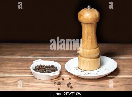 Schwarze Pfeffermühle mit Pfefferkörnern auf einer weißen Keramikplatte auf einem Holztisch und dunklem Hintergrund Stockfoto