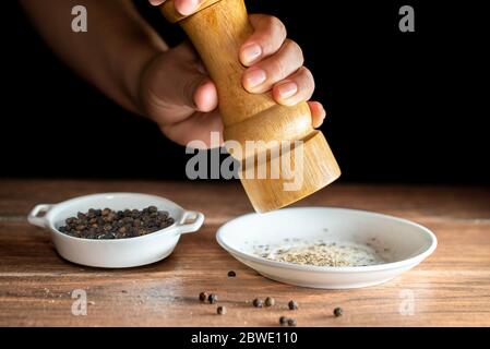 Ein Hand mahlen schwarzen Pfeffer mit einer Mühle Pfeffer und ganze Körner von schwarzem Pfeffer in einer weißen Keramikplatte auf einem Holzbrett Stockfoto