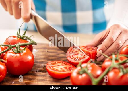 Detail der weiblichen Hände schneiden eine Tomate mit einem Messer auf einem Schneidebrett mit mehr frischen Tomaten herum Stockfoto