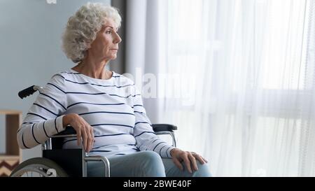 Traurige behinderte ältere Frau im Rollstuhl sitzt schaut ins Fenster Stockfoto