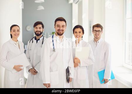 Eine Gruppe von selbstbewussten Praktizierenden Ärzten in weißen Mänteln lächelt vor dem Hintergrund der Klinik. Stockfoto