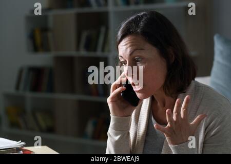 Wütende Frau mittleren Alters, die am Smartphone anruft und nachts im Wohnzimmer zu Hause sitzt Stockfoto