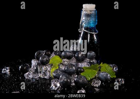 Heidelbeeren, Minzblätter, Eiswürfel und eine Flasche Brombeersaft mit Tropfen Wasser auf schwarzem Hintergrund mit Reflektion Stockfoto