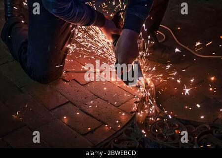 Seitenansicht der Hände eines Mannes, der an einem Metallteil einer Gartenbank arbeitet, mit einem elektrischen Schleifer, während Funken im industriellen Bereich herumfliegen Stockfoto