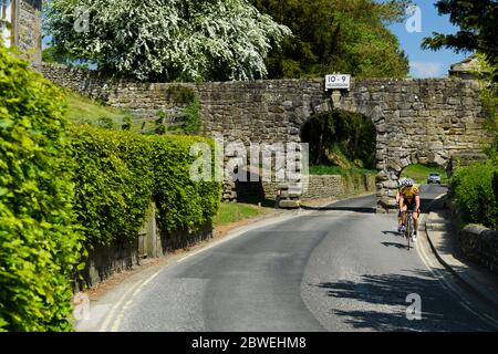 2 Radfahrer auf Landstraße durch Steinbogen (3 Bögen, Kopfraum Warnschild 10' 9'') - B6160, Bolton Abbey Village, Yorkshire, England, Großbritannien. Stockfoto
