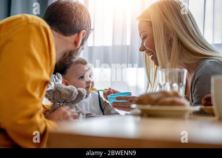 Vater und Mutter Fütterung Baby in Hochstuhl.Smiling Baby Essen in Küche.Familie, Essen, Liebe und Glück Konzept. Stockfoto