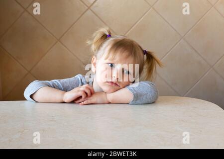 Schöne kleine Mädchen Kind traurig oder gelangweilt sitzt am Tisch in einem Zuhause Interieur. Kindheitserregungen Konzept. Quarantäne, Stockfoto