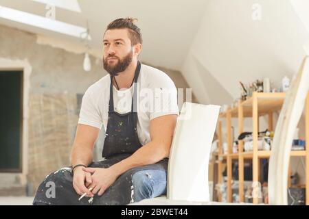 Inspirierendes Porträt eines erwachsenen bärtigen Künstlers, der nachdenklich wegschaut und raucht, während er im von Sonnenlicht beleuchteten Kunststudio auf dem Stuhl sitzt und Platz zum Kopieren bietet Stockfoto