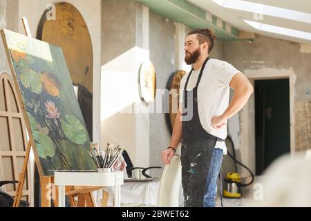 Seitenansicht Porträt von Erwachsenen bärtigen Künstler Blick auf Malerei auf Staffelei während im Loft-Art-Studio stehen, kopieren Raum Stockfoto