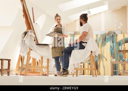 Ganzkörperportrait der weiblichen Künstlerin Malerei Bild auf Staffelei während der Arbeit in sonnenbeschienenen Kunststudio mit bärtigen Mann sitzt auf Stuhl und posiert, Kopie Stockfoto