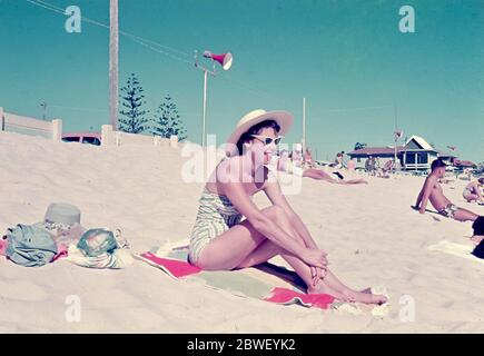 Surfers Paradise, Australien 1958: Eine junge Frau in Badekleidung, Sonnenbrille und Hut sitzt am Strand in der Sonne an der australischen Gold Coast von Queensland Stockfoto