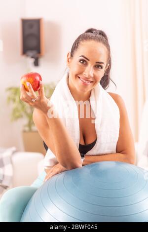 Fitness Frau, die einen Apfel essen, der sich gegen einen fitballig lehnt, nachdem sie das Heimtraining mit einem Handtuch um den Hals beendet hat. Stockfoto