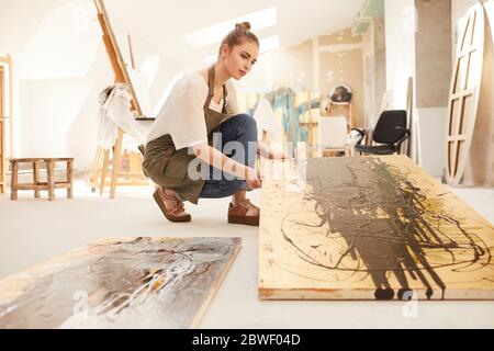 In warmen Farbtönen gehaltenes Seitenansicht Porträt einer modernen Künstlerin, die abstrakte Malerei hält, während sie im geräumigen, von Sonnenlicht beleuchteten Kunststudio auf dem Boden sitzt, Kopie sp Stockfoto