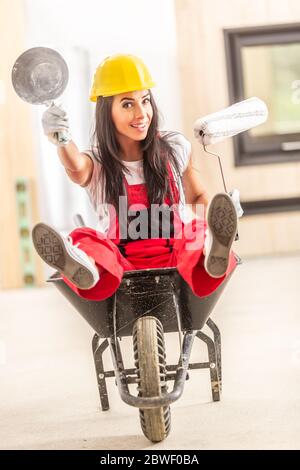 Schönes Mädchen sitzt in einer Schubkarre innerhalb der Baustelle mit Bauwerkzeugen in den Händen tragen Overall und einen Sicherheitshelm. Stockfoto