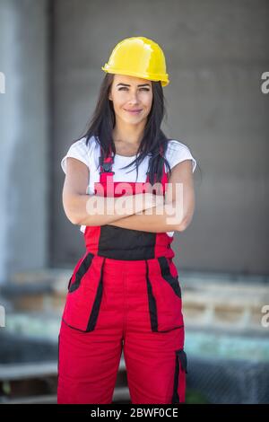 Die Baumeisterin steht mit gekreuzten Armen auf einer Baustelle, die in Arbeitsoveralls und einem gelben Hut gekleidet ist. Stockfoto