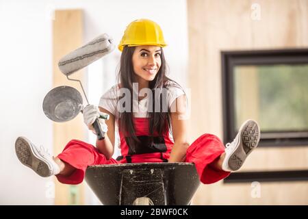 Schönes Mädchen sitzt in einer Schubkarre innerhalb der Baustelle mit Bauwerkzeugen in den Händen tragen insgesamt und einen harten Hut. Stockfoto