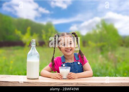 Schöne kleine Brünette Mädchen sitzt an einem Tisch, hält ein Glas Milch und lächelt, eine Flasche Milch steht daneben, gegen ein grünes Feld und blau s Stockfoto