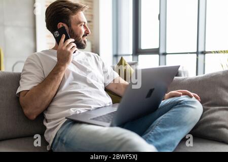 Junger Mann, der einen Laptop benutzt, während er am Telefon auf der Couch sitzt Stockfoto