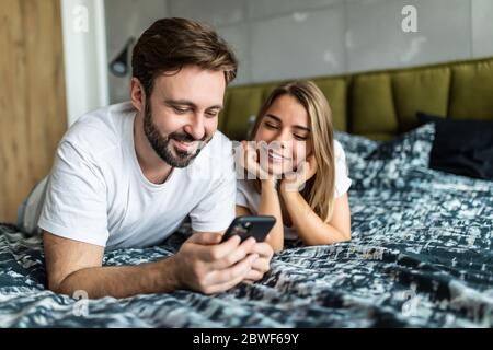 Lächelndes Paar mit Smartphone, das zusammen auf dem Bett liegt. Happy Freund und Freundin Check Social Networks Nachrichten am Morgen. Stockfoto