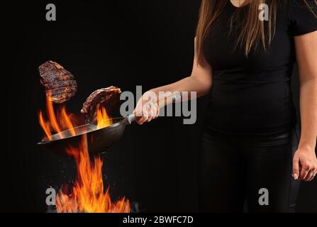 Brunette Koch mit tätowierten Händen, gekleidet in Leggings und T-Shirt. Wok-Pfanne über Feuer halten und zwei Rindersteaks vor schwarzem Hintergrund braten. Co Stockfoto