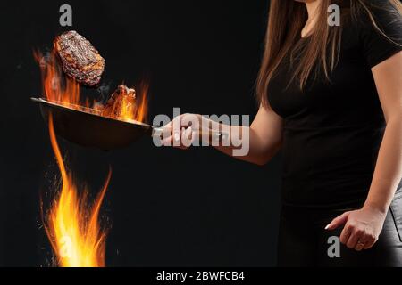 Frau mit tätowierten Händen, gekleidet in Leggings und T-Shirt. Halten brennende Wok Pfanne über Feuer und Braten zwei Rindfleisch Steaks, schwarzer Hintergrund. Seitenansicht Stockfoto