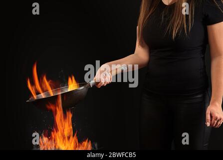 Brunette Dame mit tätowierten Händen und langen Haaren, gekleidet in Leggings und T-Shirt. Hält eine Wok Pfanne über Feuer vor schwarzem Studio Hintergrund. Kochen Stockfoto