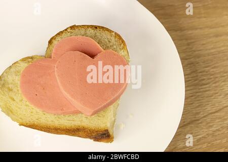 Romantisches Frühstück am Valentinstag: Weißbrot-Sandwich mit Schinken in Herzform auf Holzhintergrund. Valentinstagsmenü. Wurst er Stockfoto