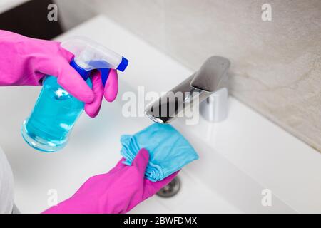 Weibliche Hände tragen Gummihandschuhe halten Mikrofasertuch und Sprühflasche Desinfektionsmittel zu desinfizieren Waschbecken Wasserhahn im Bad. Stockfoto