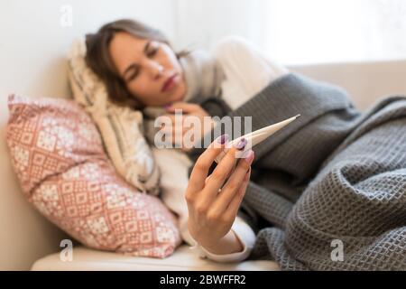 Kranke junge kaukasische Frau liegt auf dem Sofa, in Schal gewickelt, unter dem Deckeltuch, Grippesymptome, Halten und Betragen Thermometer, konzentrieren sich auf sie Stockfoto