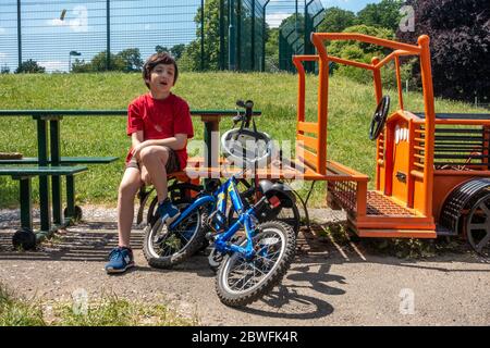 Ein kleiner Junge sitzt auf einer Bank in einem Park, um sich auszuruhen. Er hat sein Fahrrad dabei. ES ist ein warmer Sommertag. Stockfoto