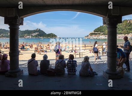 Eine Gruppe von Menschen, die an einem sonnigen Tag aus einem Tierheim über dem überfüllten Strand von San Sebastian blicken. Stockfoto