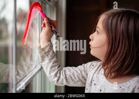 Ein kleines ernstes Kind zeichnet einen Regenbogen auf ein Fenster Mit roter Markierung Stockfoto