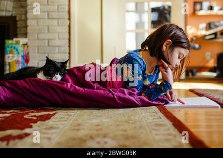 Ein Kind rollt sich im Schlafsack auf dem Boden auf Mit Katze macht Schularbeiten Stockfoto