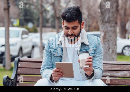 Ein bärtiger, stilvoll gekleideter Inder sitzt auf einer Bank und nutzt sein Tablet. Stockfoto