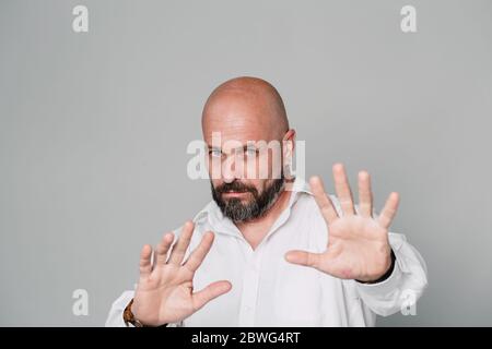 Ein gutaussehender Mann mittleren Alters in einem weißen Hemd ist verärgert und hält seine Hände vor der Kamera, als ob er sich vor etwas auf einem grauen Hintergrund versteckt Stockfoto