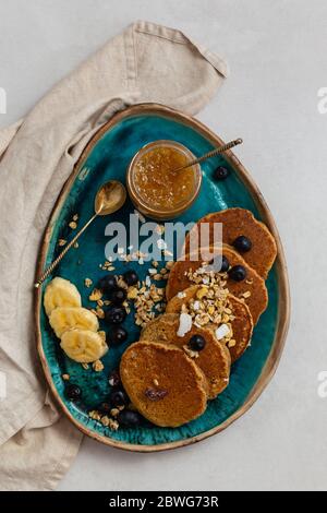 Obere Ansicht der Haferflocken Banana Pfannkuchen serviert mit Heidelbeeren, Scheiben von Banane und Zitrone Marmeladein ein Glas in einem türkisfarbenen Keramik pla platziert Stockfoto