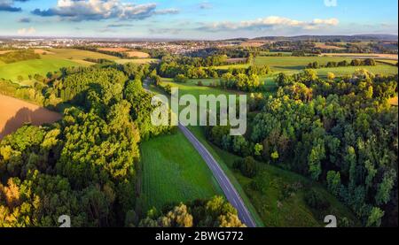 Idyllische deutsche Landschaft, aufgenommen von oben bei frühmorgendlicher Sonneneinstrahlung: Wiesen und Wälder mit blauem Himmel und eine Straße, die zu einer kleinen Stadt am Horizont führt Stockfoto