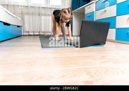 Ein kleines Mädchen in einem schwarzen Turntrikot macht Gymnastik zu Hause online vor einem Laptop. Platz für Text und Kopierer. Stockfoto