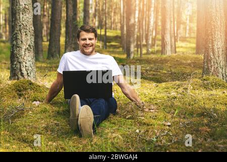 Junger lächelnder Mann, der im Moos sitzt und Laptop im Wald benutzt Stockfoto