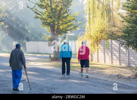 Alberton, Südafrika - ein unidentifizierter behinderter alter Mann und zwei fitt unidentifizierte Wanderer isoliert auf der gleichen Straße in der frühen Morgensonne Stockfoto