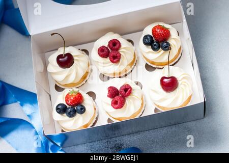 Cupcakes mit Schlagsahne, frischen Kirschen, Himbeeren, Heidelbeeren, Erdbeeren in Geschenkbox mit blauem Band. Stockfoto