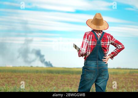 Besorgte Landwirtin mit digitalen Tablet im Maisfeld Blick auf schwarzen Rauch am Horizont, Konzept der Versicherung in der Landwirtschaft und Landwirtschaft Stockfoto