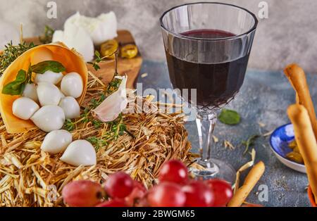 Baby Mozzarella Käse auf einem Heu mit Knoblauch und Trauben, ein Glas Rotwein auf blauem Hintergrund. Stockfoto