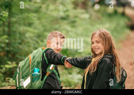 Happy boy und Mädchen gehen Wandern mit Rucksäcken auf Waldstraße hellen sonnigen Tag. Der Junge mit seiner Schwester geht wandern. Stockfoto