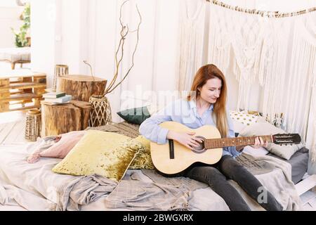 Eine Erwachsene Frau spielt die Gitarre auf dem Bett in einem Boho-Stil Zimmer sitzen. Schöne und gemütliche Atmosphäre. Stockfoto