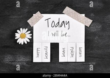 Weiße abreißende Stub-Note mit Text 'Today I will be...' und Worten 'glücklich, schön, mutig, hilfsbereit' und 'Strong' auf dunkelschwarzem Hintergrund mit Gänseblümchen Stockfoto